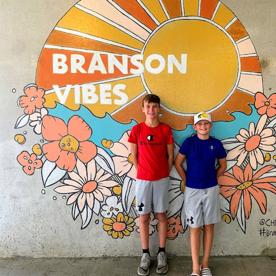 Two boys stand in front of a mural that reads,  'Branson Vibes'.