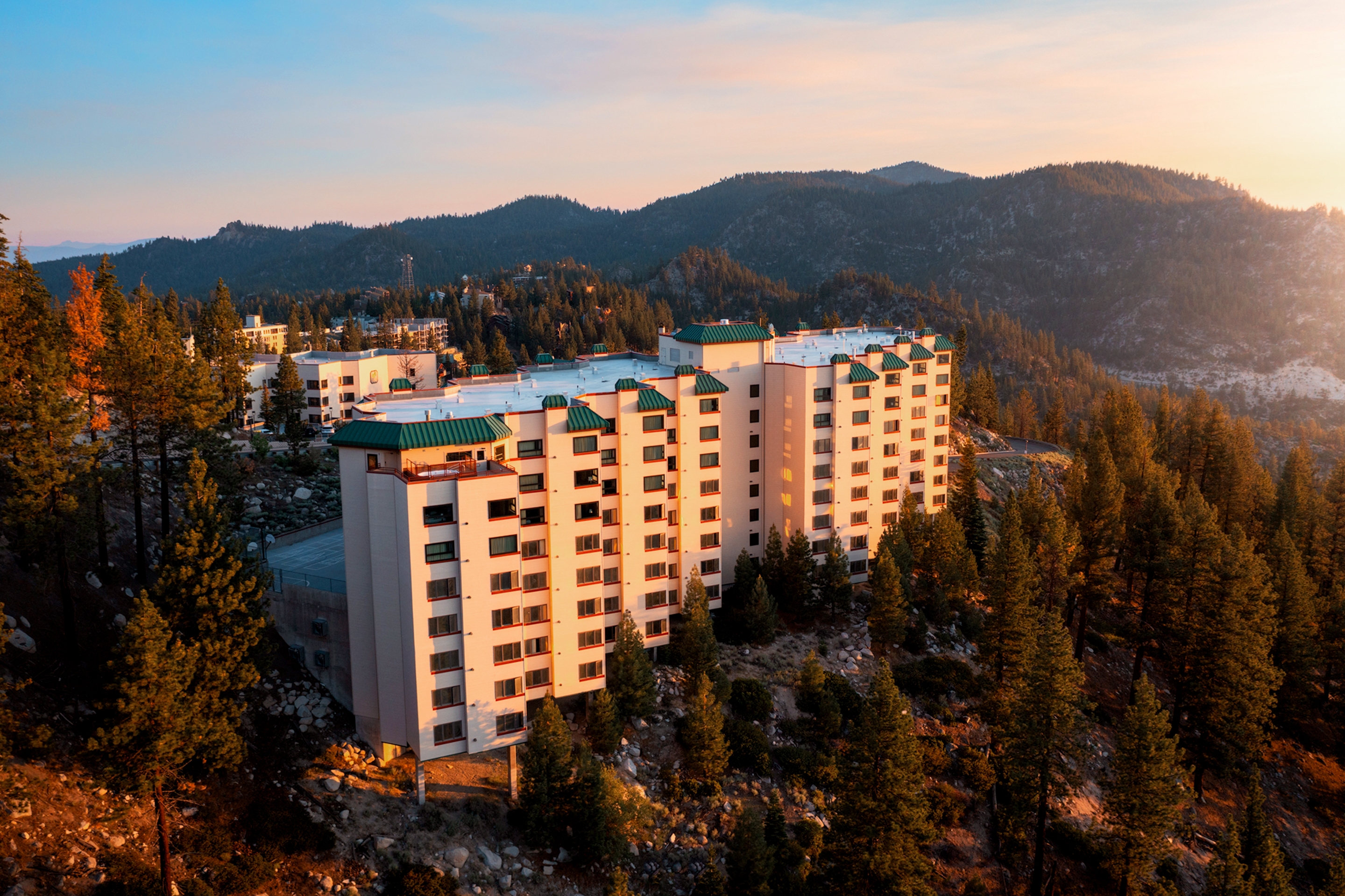 A drone view of our Tahoe Ridge Resort exterior located in Lake Tahoe, Nevada surrounded by pine trees of various heights and the mountain ridge as the sun sets.