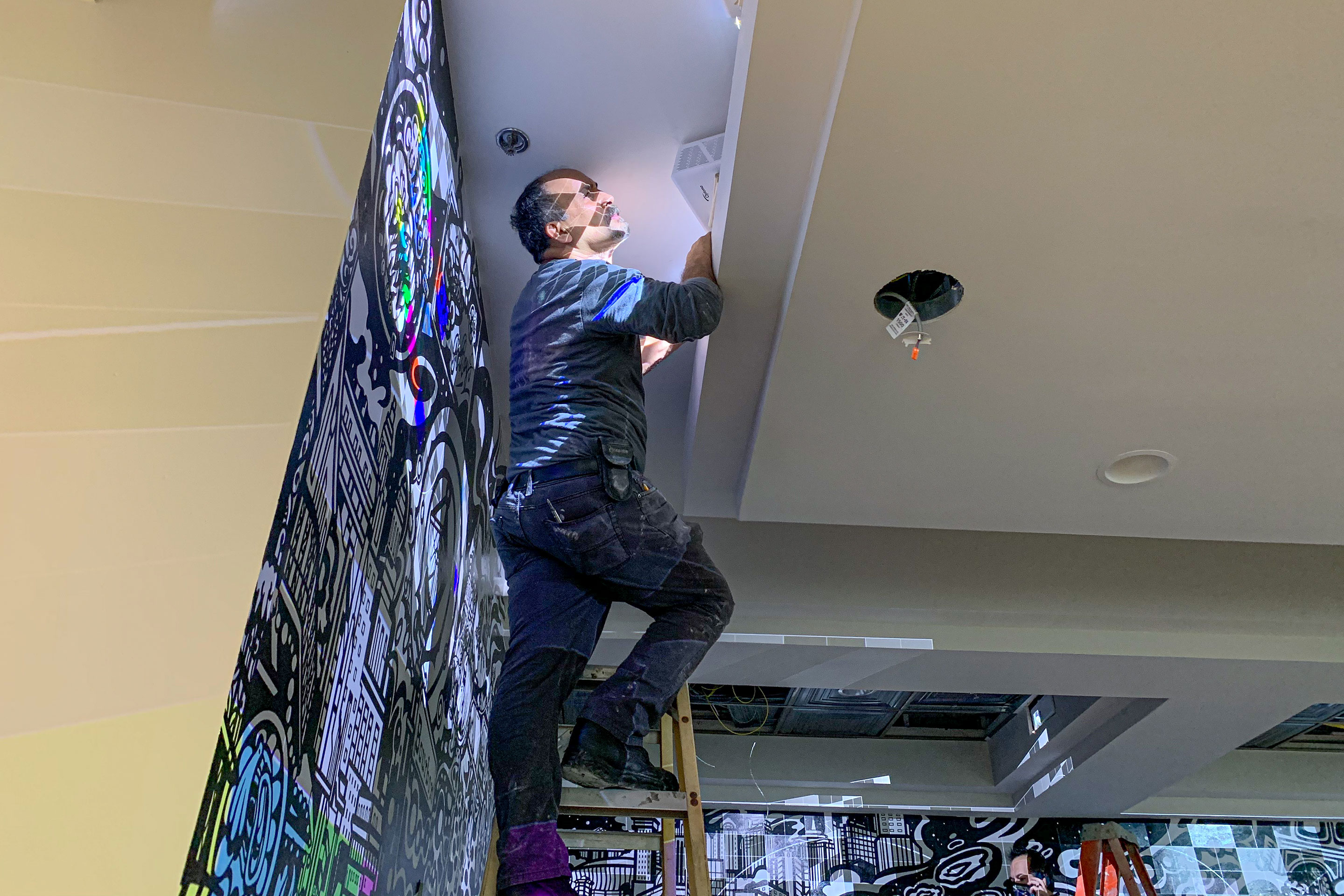 A man climbing a ladder while adjusting colored projectors for the outlined mural art featured in the lobby of our resort in New Orleans, Louisiana.
