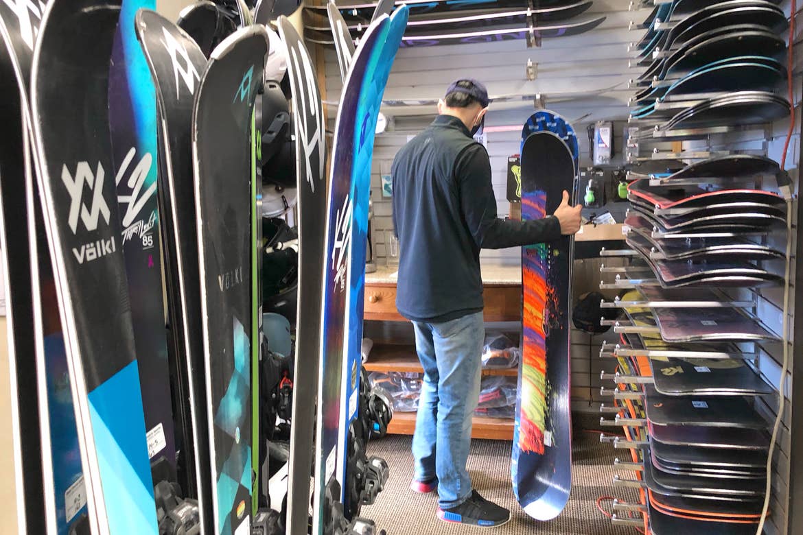 Team Member, 'Ernie', prepares to wax a snowboard in the Resort Sport Shop surrounded by various snowboards.