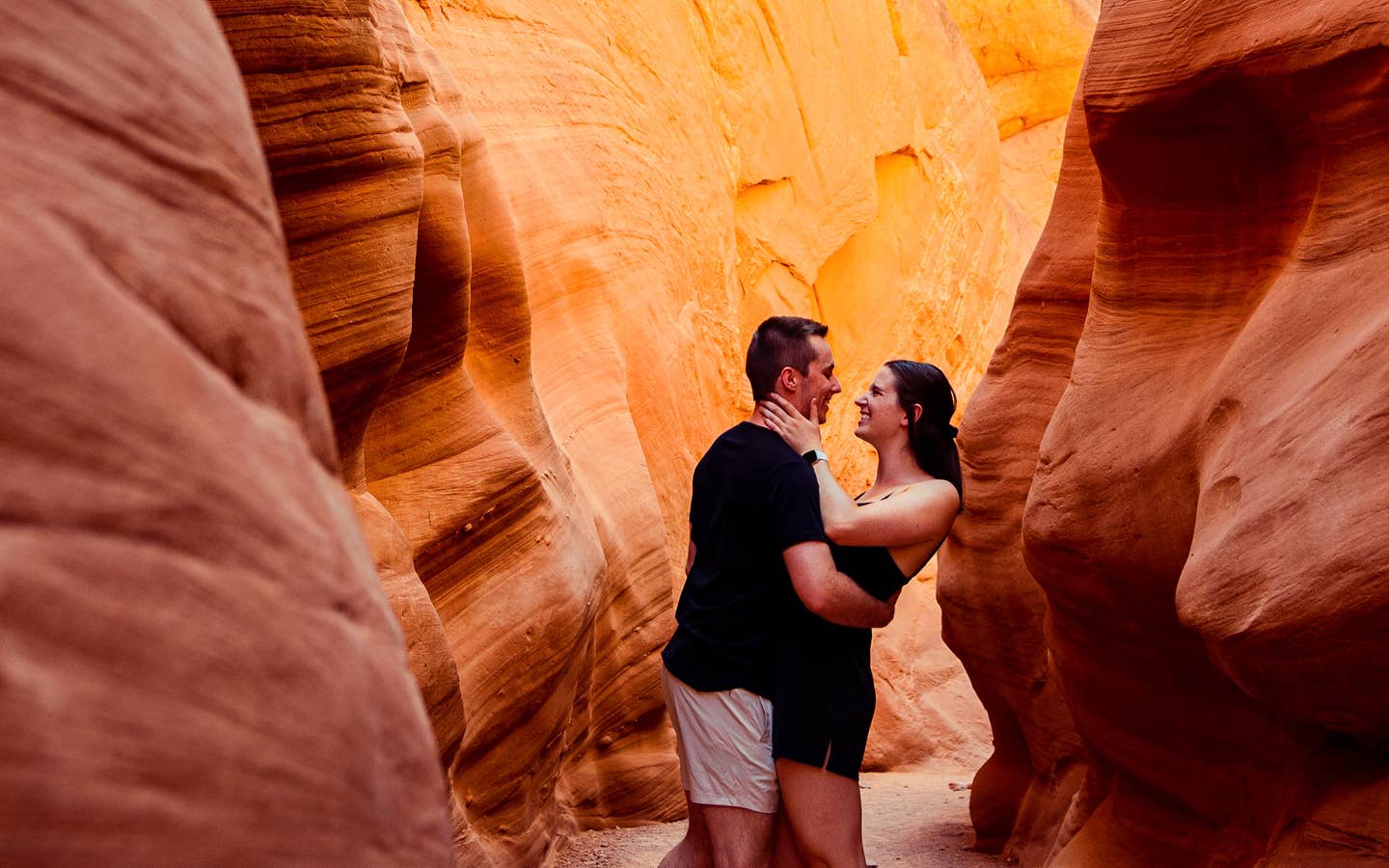 A man and woman wearing black outfits embrace in a red rock formation.