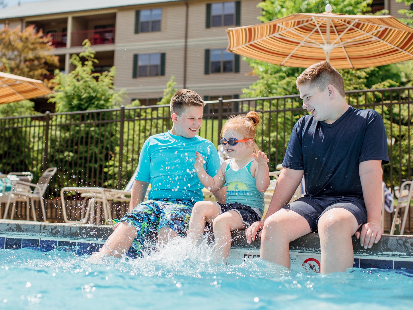 Two young caucasian tween boys (left and right) wearing swim shirts and shorts sit with their feet in a pool with a young caucasian girl (middle) wearing a swimsuit, swimming shorts and goggles as she splashes her feet.