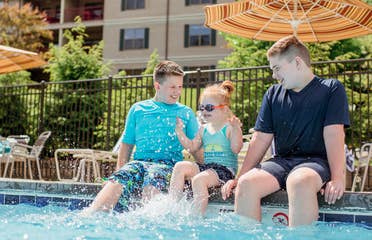 Two young caucasian tween boys (left and right) wearing swim shirts and shorts sit with their feet in a pool with a young caucasian girl (middle) wearing a swimsuit, swimming shorts and goggles as she splashes her feet.