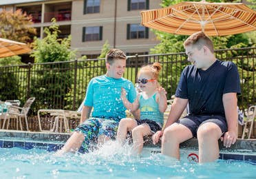 Two young caucasian tween boys (left and right) wearing swim shirts and shorts sit with their feet in a pool with a young caucasian girl (middle) wearing a swimsuit, swimming shorts and goggles as she splashes her feet.