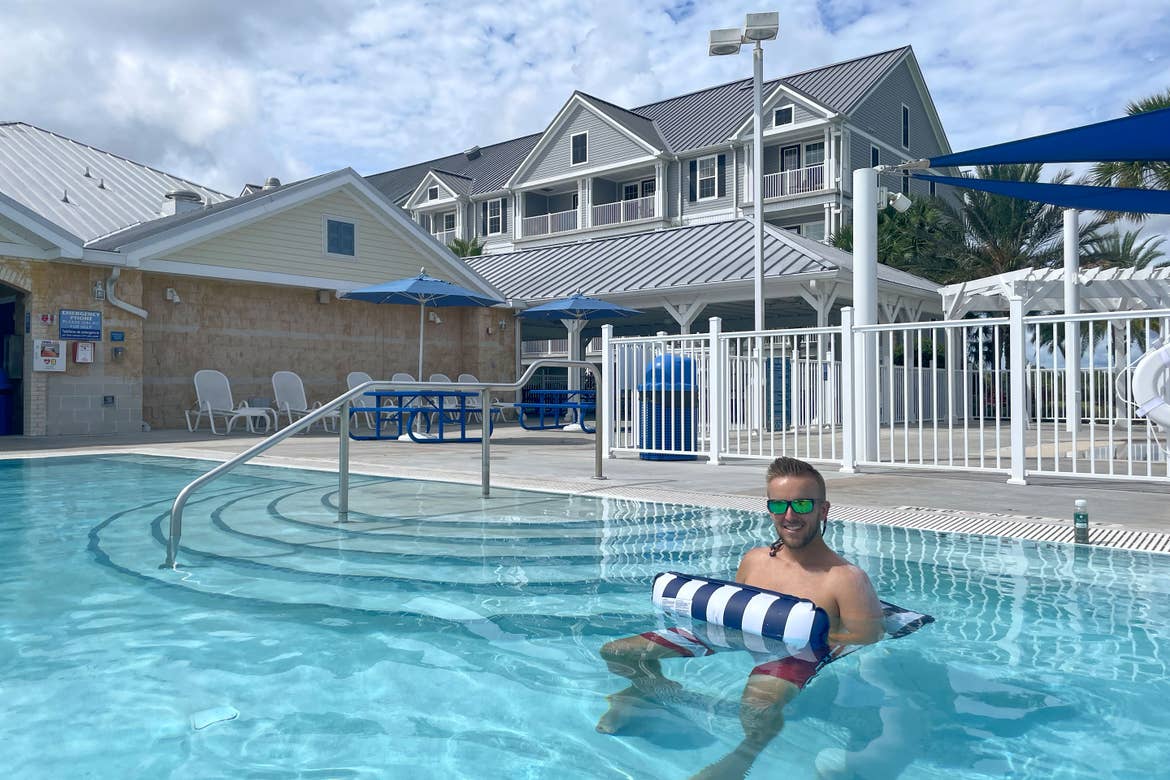 A man wades in an outdoor pool on a blue and white striped inflatable near a resort building.