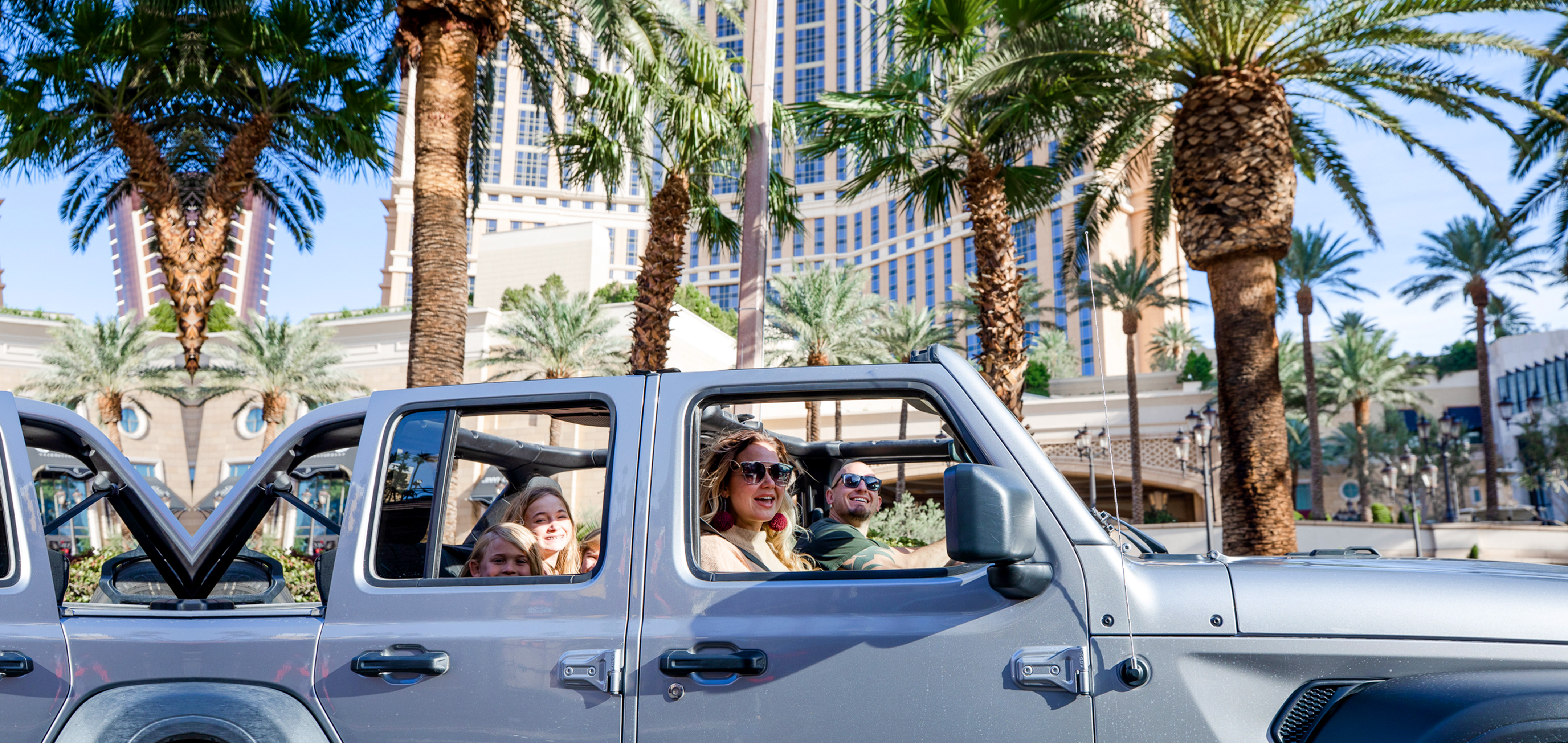 A man drives a silver Jeep Wrangler with one female passenger and three children in the back though the Las Vegas strip with the roof shell off.