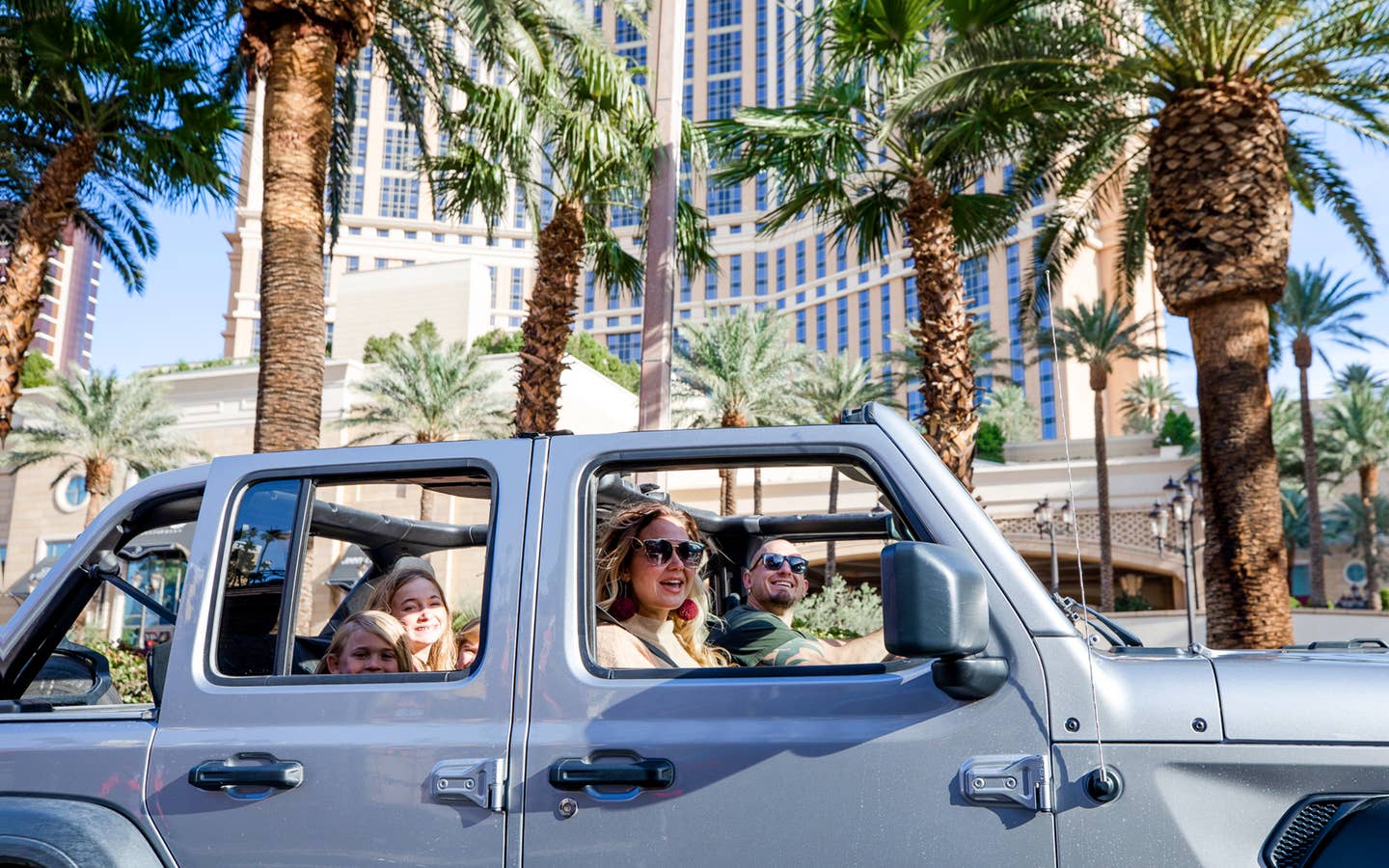 A man drives a silver Jeep Wrangler with one female passenger and three children in the back though the Las Vegas strip with the roof shell off.