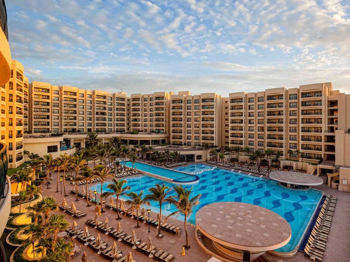 Outdoor pool surrounded by palm trees, sun chairs and resort buildings at sunset in Mexico.