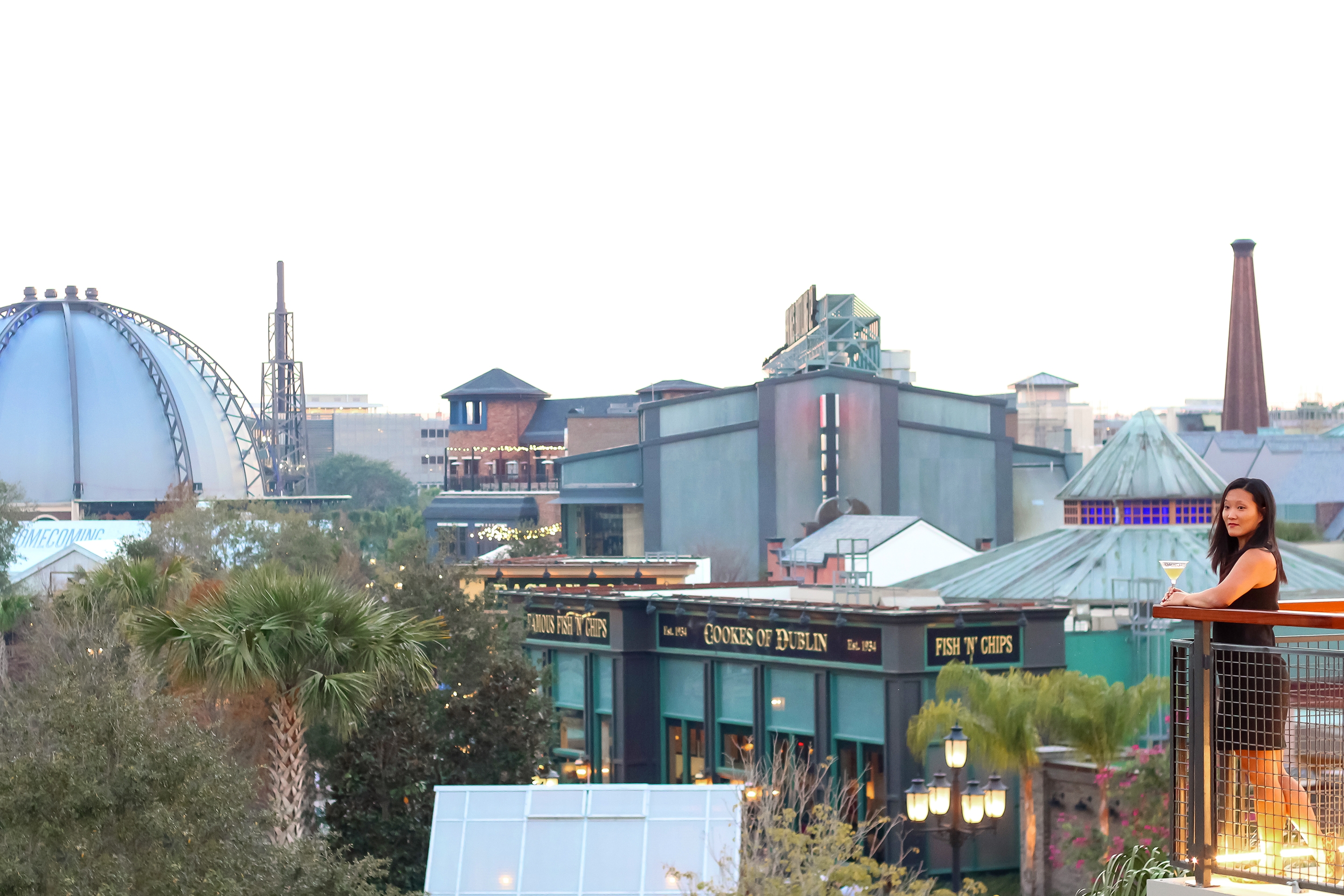 An Asian woman wearing a black dress stands on a balcony overlooking the scenery of Disney Springs.
