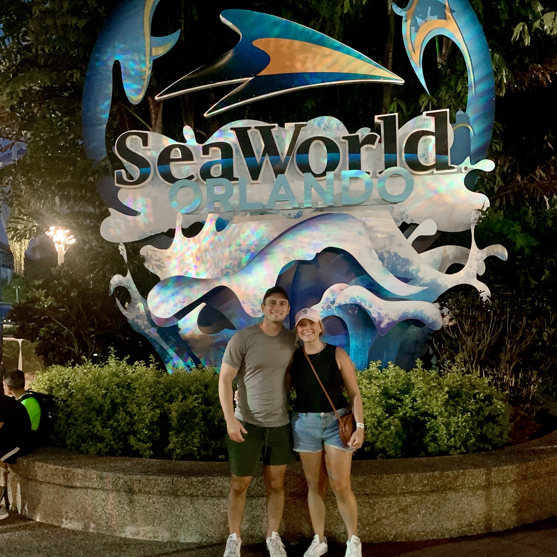 A Caucasian male wearing a grey t-shirt, black cap (right) and a Caucasian female wearing a pink baseball cap and black tank top stand near the SeaWorld Orlando sign icon.