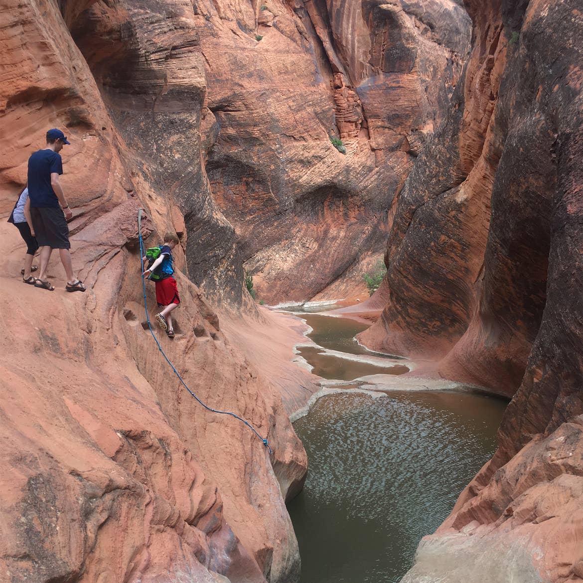 A many and two young boys wearing basketball shorts, sandals and backpacks walk on a rock formation trail on orange rocks at Zion National Park near a ravine.