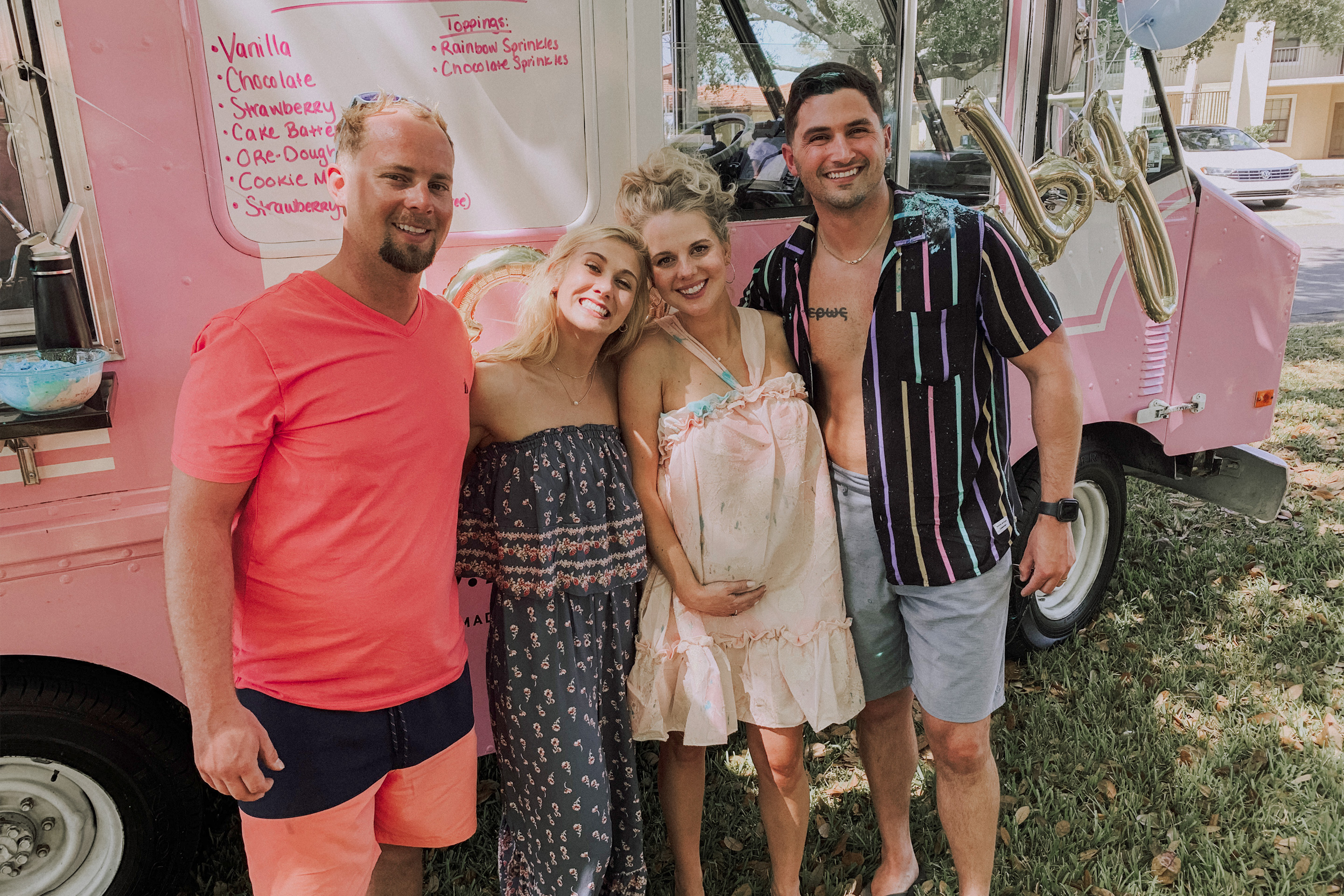 A caucasian woman wearing a pink dress (middle-right) and Latinx male wearing a striped black button-down shirt, and light shorts (right) stand near another caucasian couple (left) in front of a pink food truck.