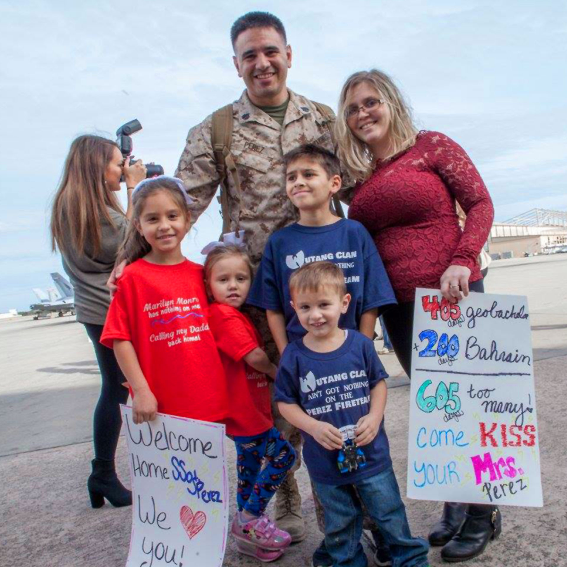 Featured Member, Sara Perez (top-right), stands with her husband in his Marine uniform and their four children holding signs for his Homecoming.