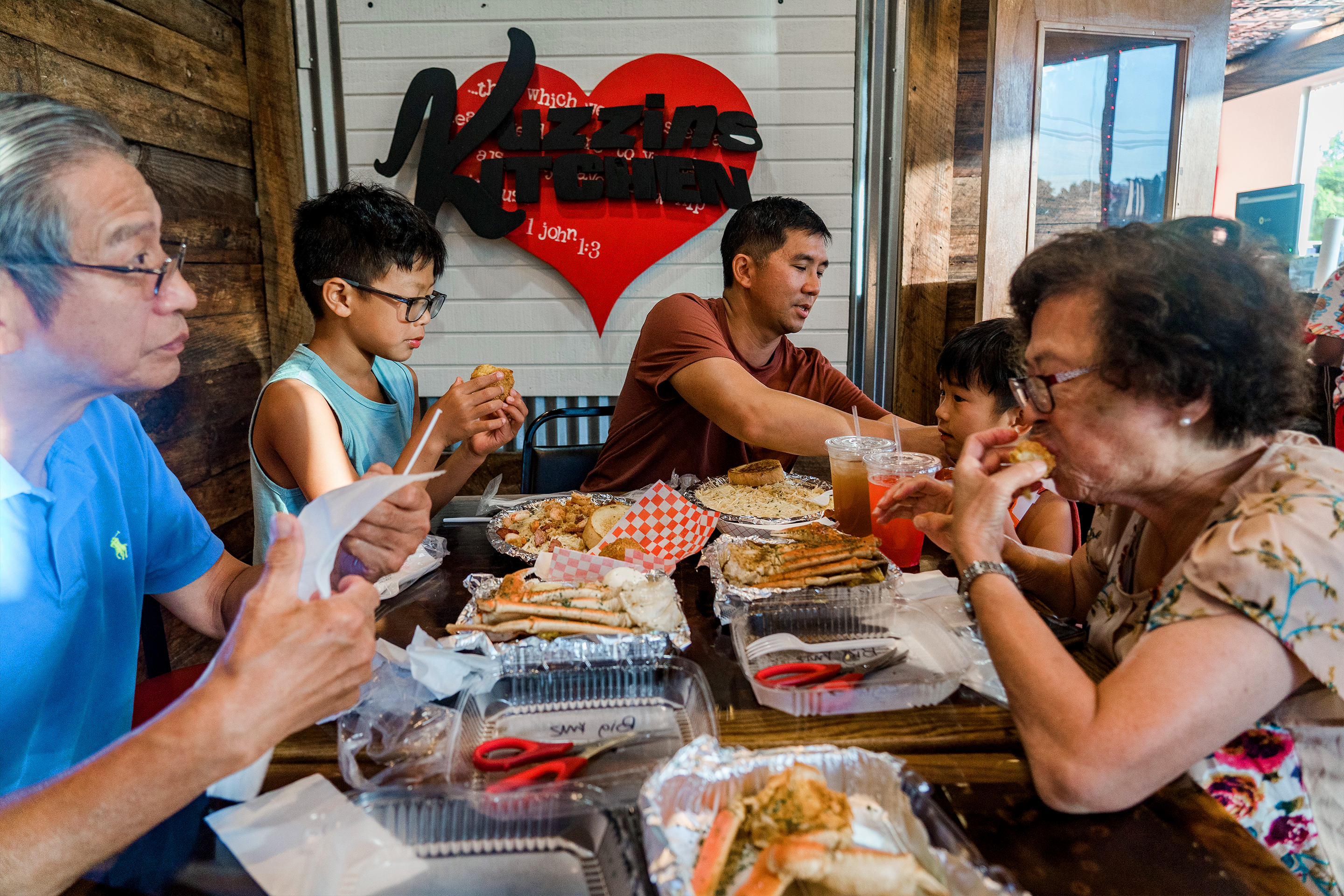A multi-generational Asian family enjoys various food near a shiplapped wall with a heart-shaped marquee that reads 'Kuzzin's Kitchen'