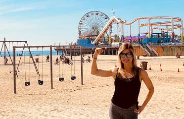 Featured Co-author, Christine, stands on Venice Beach wearing black tanks and leggings in front of the boardwalk and various exercise areas.
