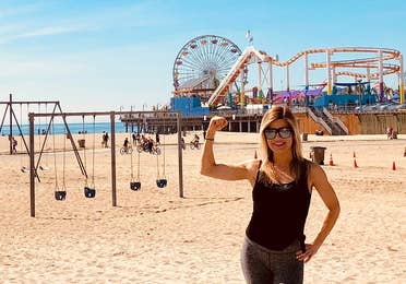 Featured Co-author, Christine, stands on Venice Beach wearing black tanks and leggings in front of the boardwalk and various exercise areas.