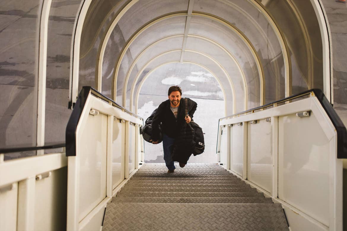 Sara's husband is running up a covered staircase at the airport carrying his luggage and smiling.