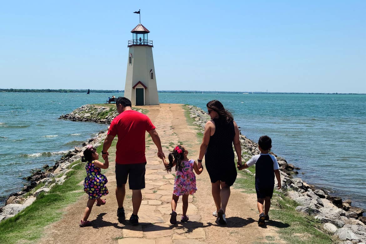 Three toddlers walk with a man and a woman down a pier to a lighthouse.