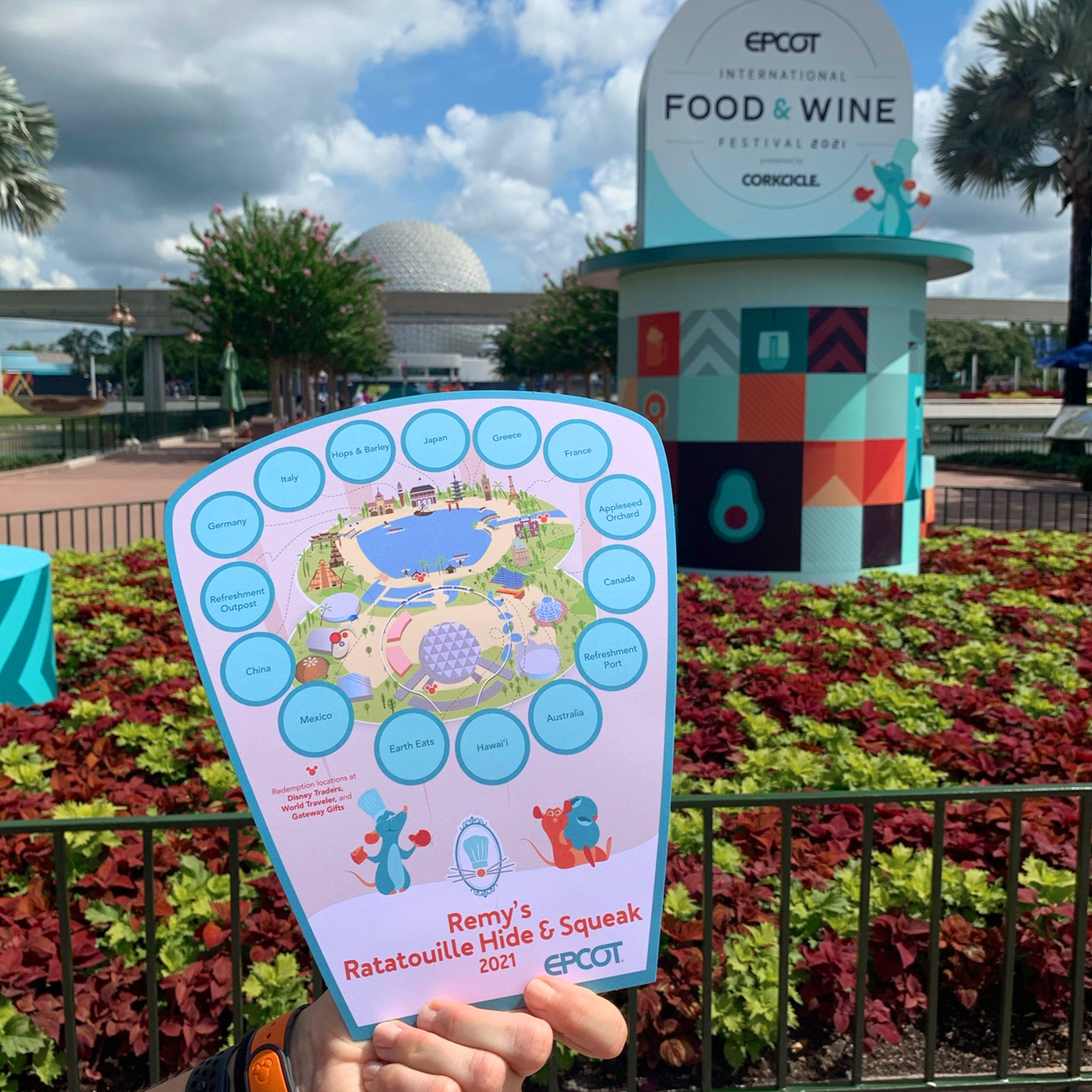 A hand holds a map for 'Remy's Hide & Squeak Scavenger Hunt' near a garden display sign with the Epcot Geosphere in the background.