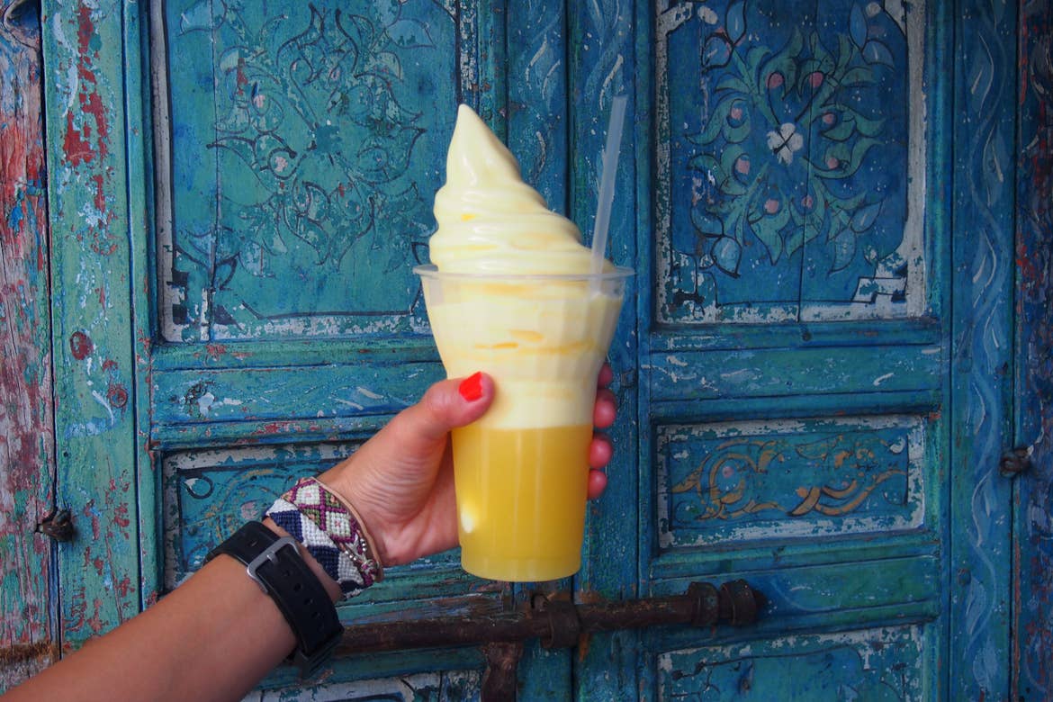 A woman's hand holds a citrus-yellow Dole Whip float in front of a blue distressed door at Walt Disney World Resort.