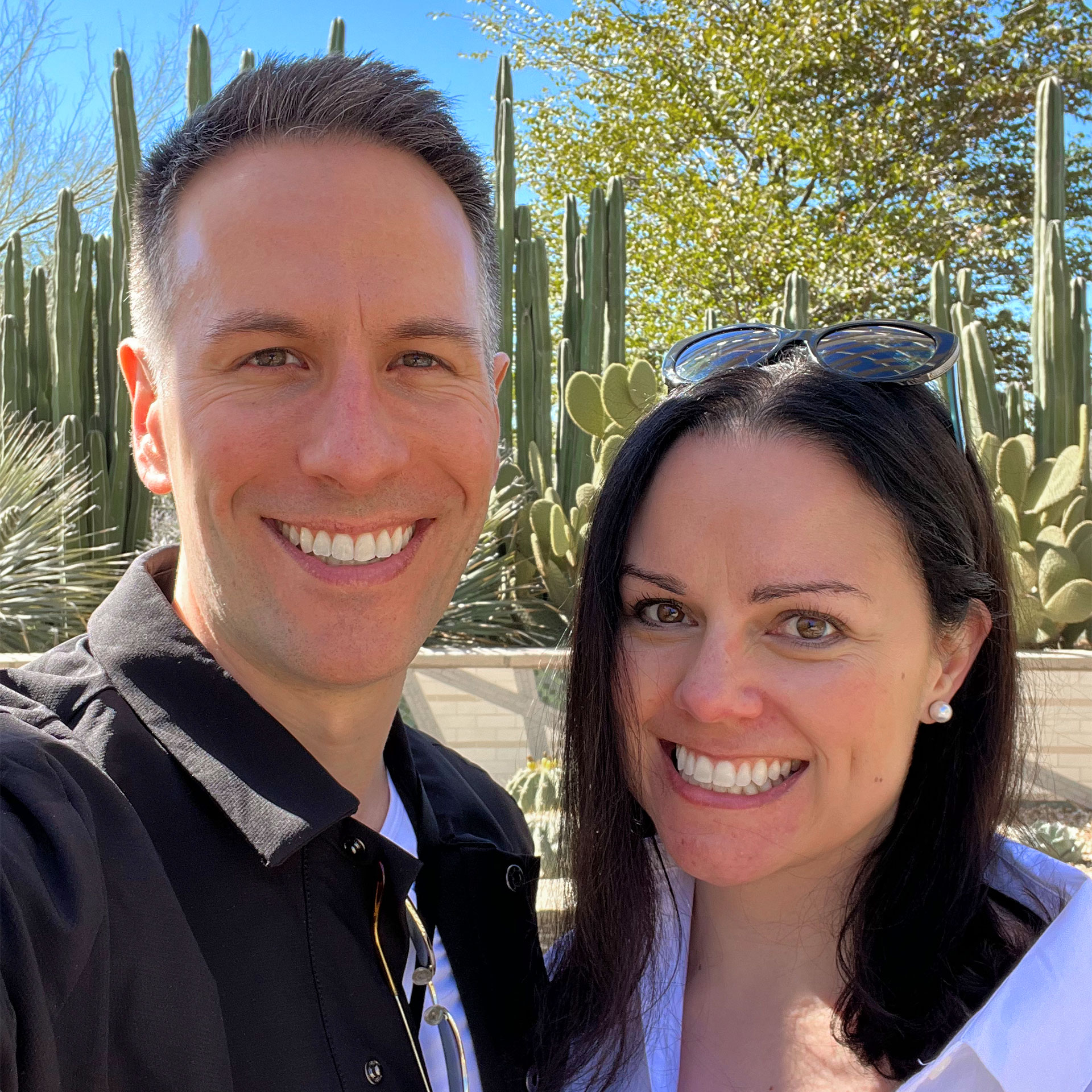 A man in a black jacket (left) and a woman in a white jacket and sunglasses (right) stand in front of cacti outdoors.