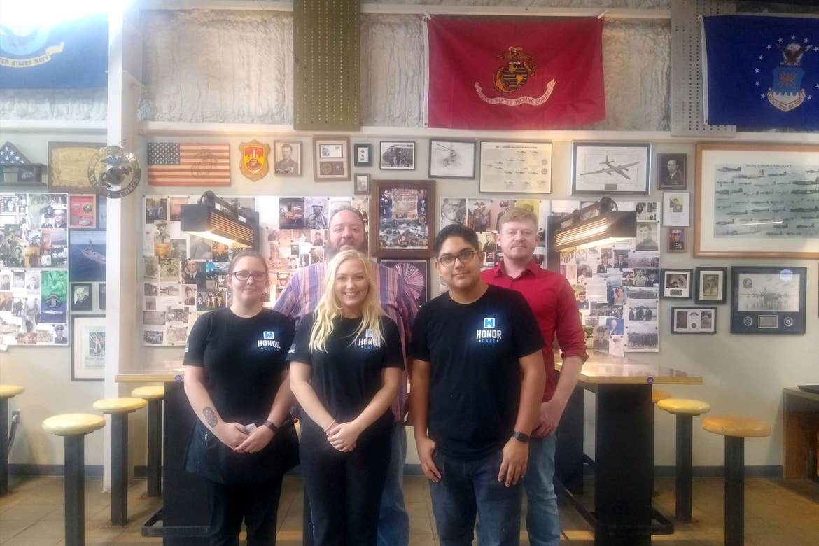 Several servers dressed in Jeans and black shirts, or red button-ups stand in front of a wall decorated with pictured of Veterans and military flags and memorabilia.