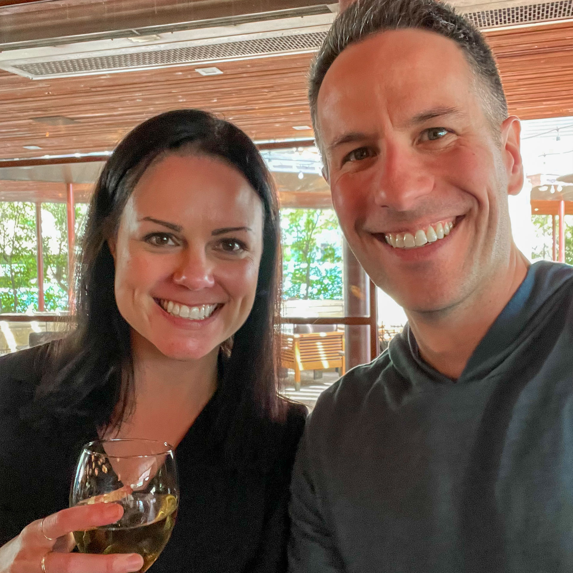 A woman in a black sweater (left) holds a glass of white wine next to a man in grey hoodie (right) on a terrace.