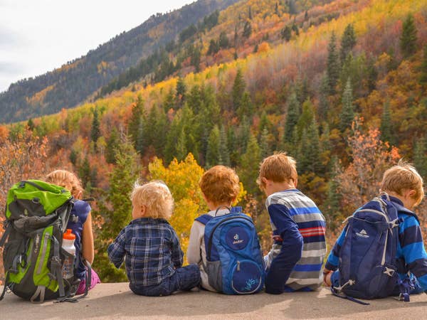 Five caucasian children (left to right: a girl, and four boys) wear backpacks and patterned shirts while sitting on ledge looking at colorful fall foliage.