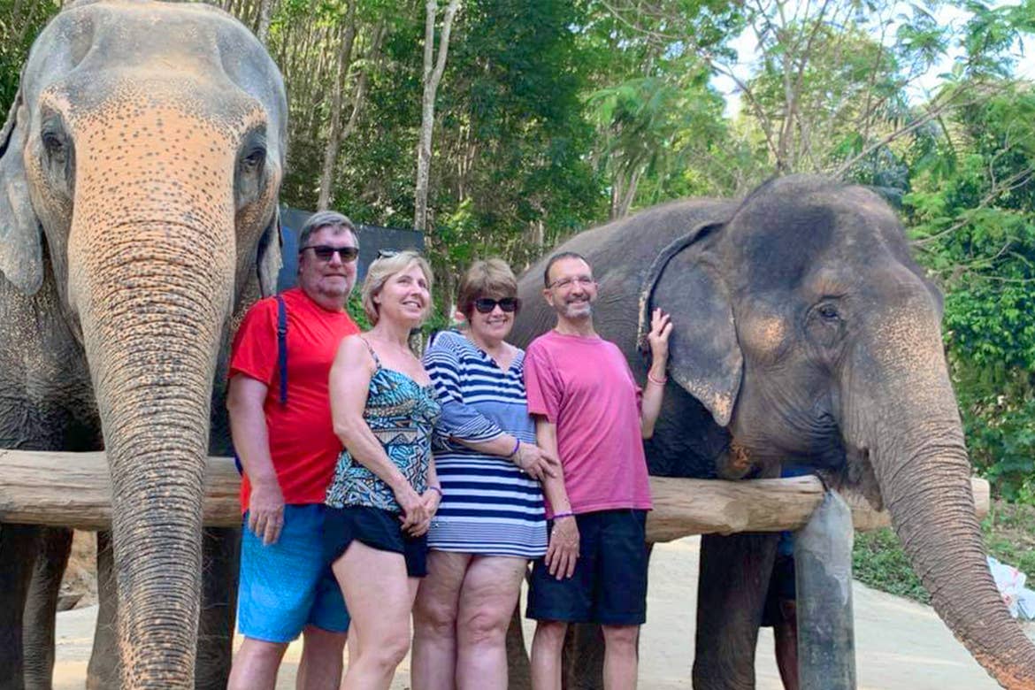 Two caucasian men and two women stand in front of two elephants near a wooden fence and jungle foliage in Phuket, Thailand.