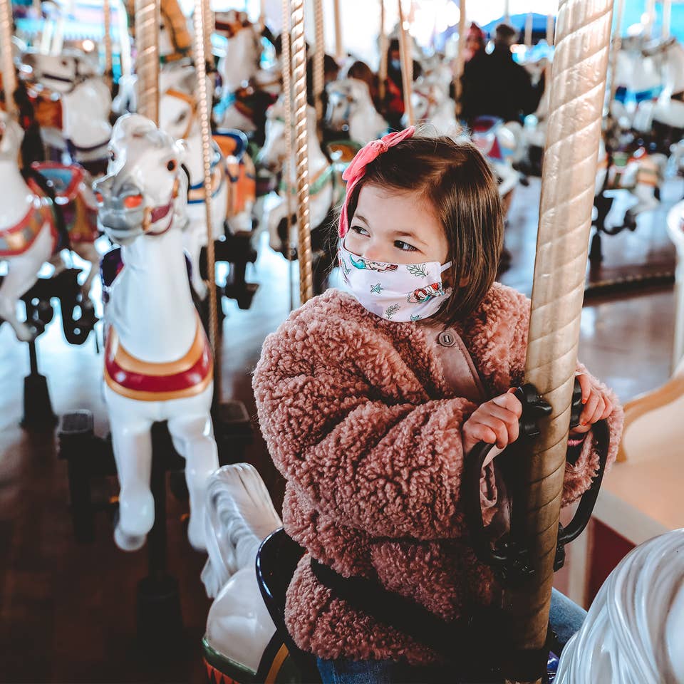 Roux rides a white horse on Prince Charming Regal Carrousel at Magic Kingdom Park at Walt Disney World® Resort wearing a pink fuzzy jacket and a 'Little Mermaid' face mask.