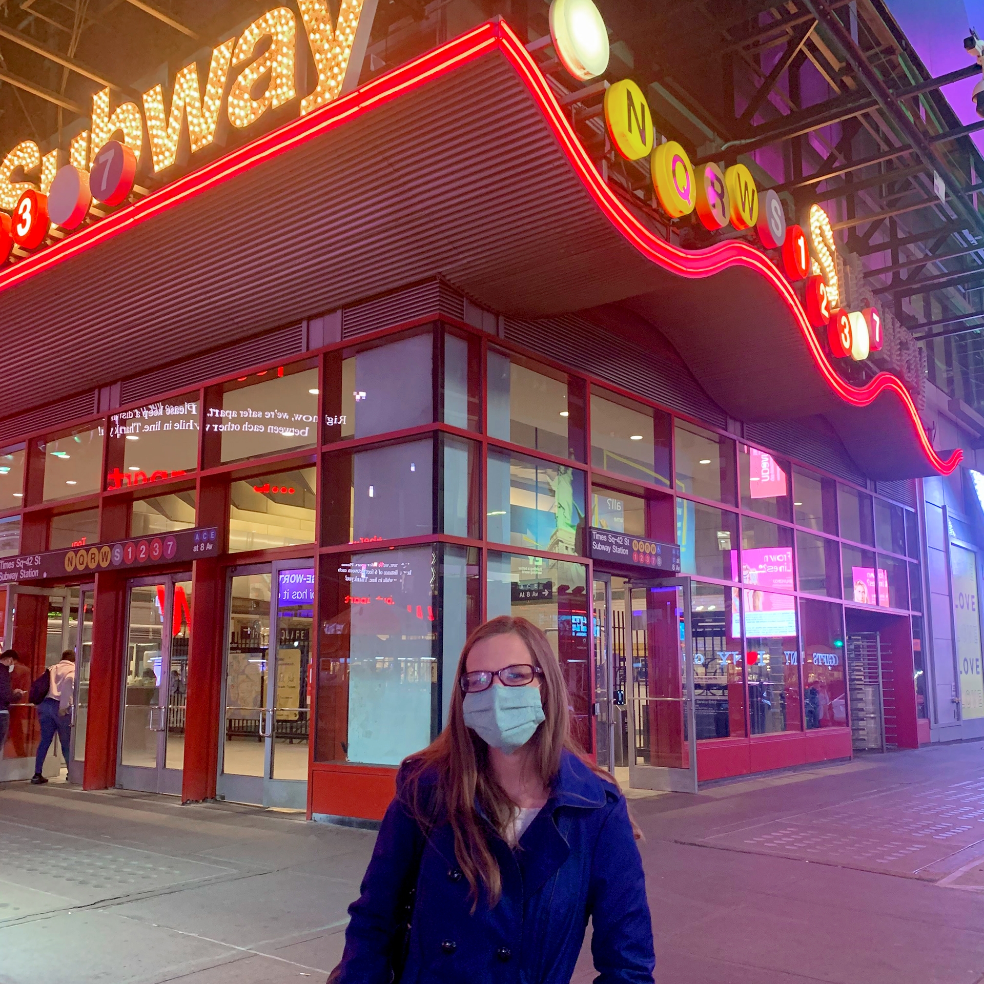 A Caucasian woman with long hair wears a pair of glasses, a safety mask and a jacket outside of the Times Square subway station in New York City at night.