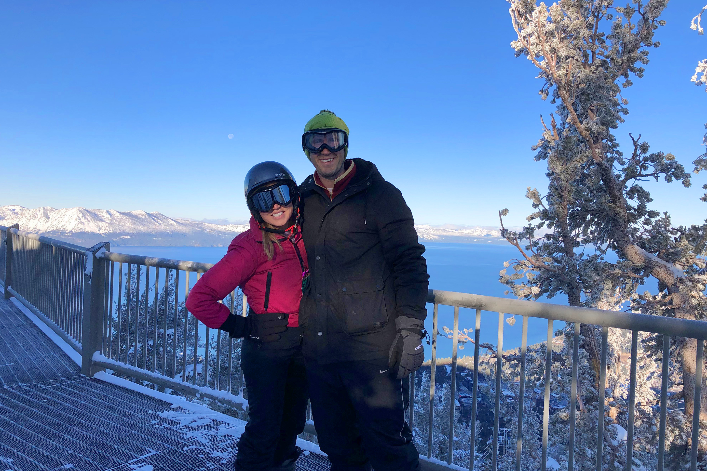 A woman (left) wearing a pink snowboard jacket, black snow pants, and a helmet with goggles stands near a man (right) wearing goggles, a green beanie, and black snowboarding jacket and black snow pants.