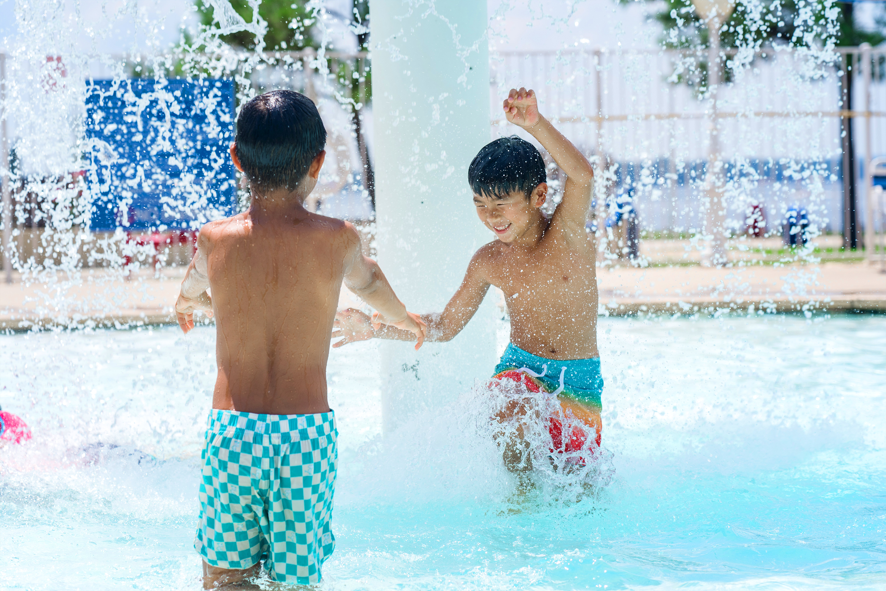 Two young, Asian boys wear multicolored swim trunks while playing under an outdoor pool fountain.
