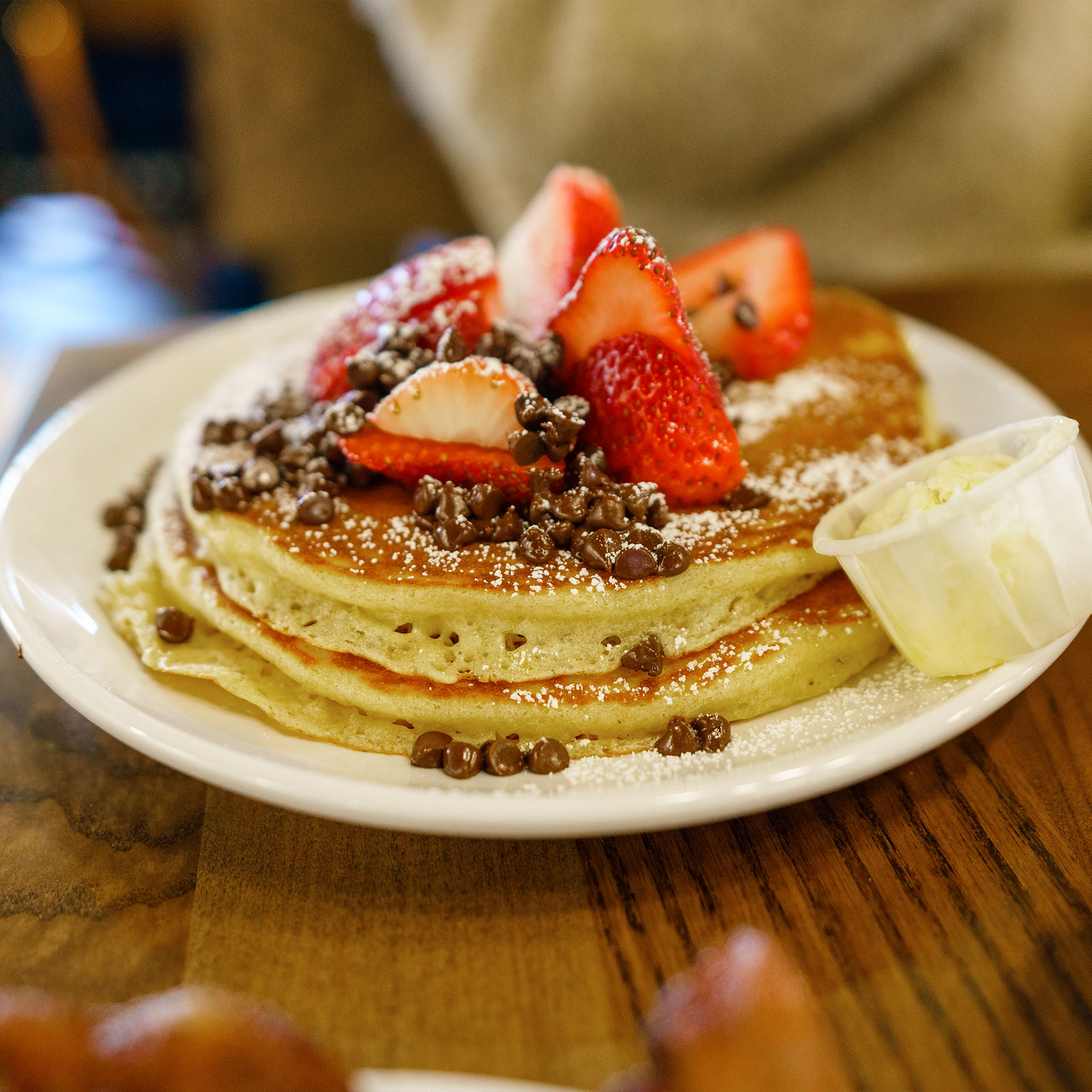 A short stack of pancakes topped with powdered sugar, pecans and fruit is placed on a wooden dining table.