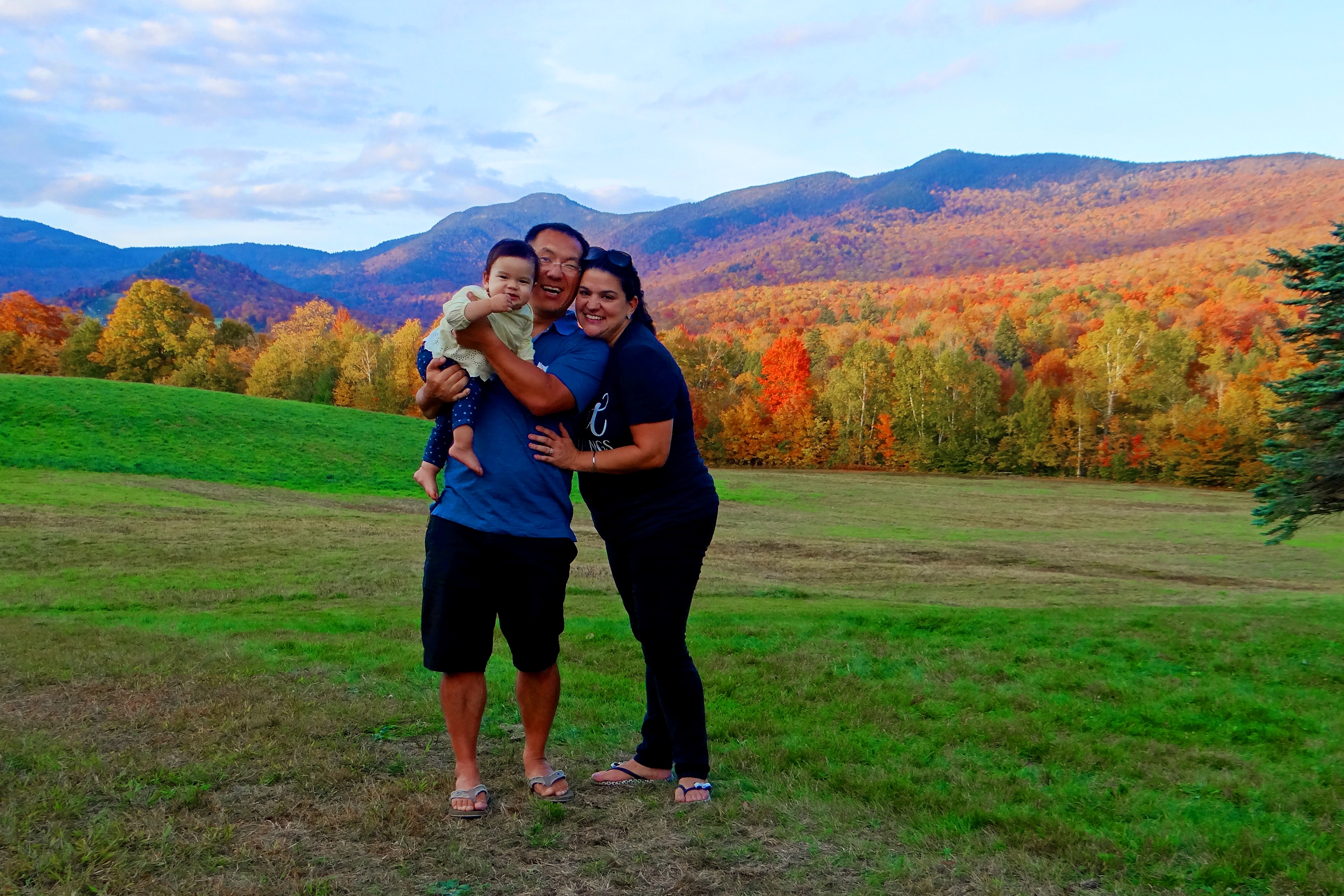 An Asian Pacific Islander toddler (left) is held by an Asian Pacific Islander man (middle) standing near a woman (right) surrounded by lush fall foliage.