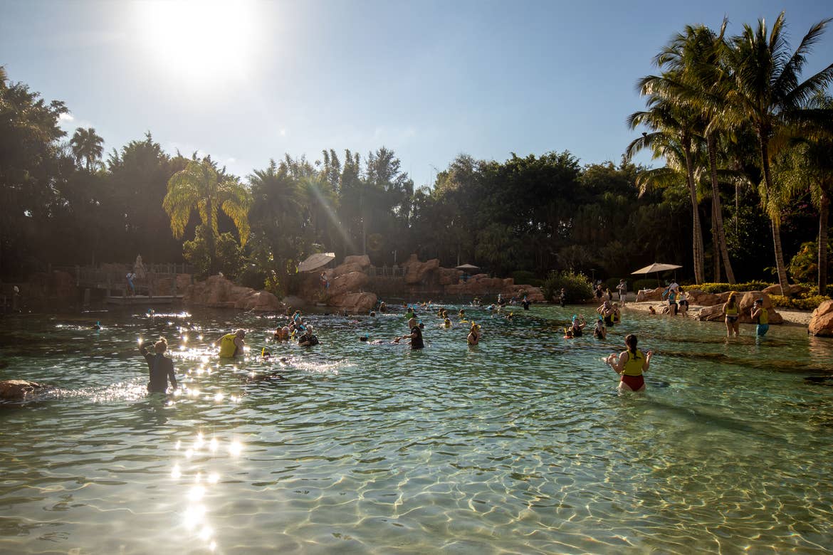 The sun shines on an outdoor water area containing tens of guests in swimwear and lifejackets.