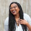 Krystin Godfrey wears a white blouse, braids and Tortoise shell glasses in front of a cement wall.