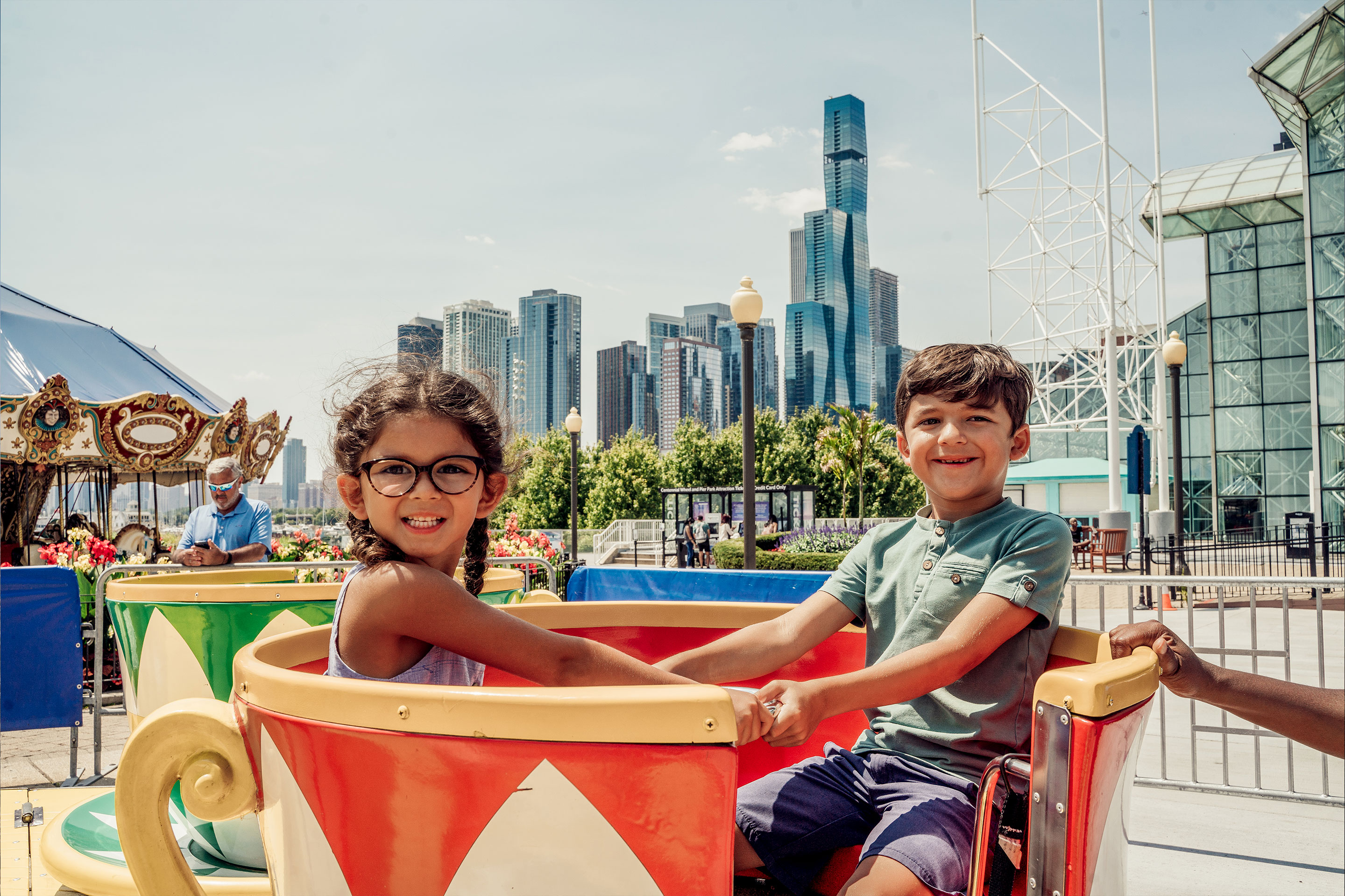 A young girl with glasses (left) and a young boy (right) sit in a spinning teacup ride outdoors.
