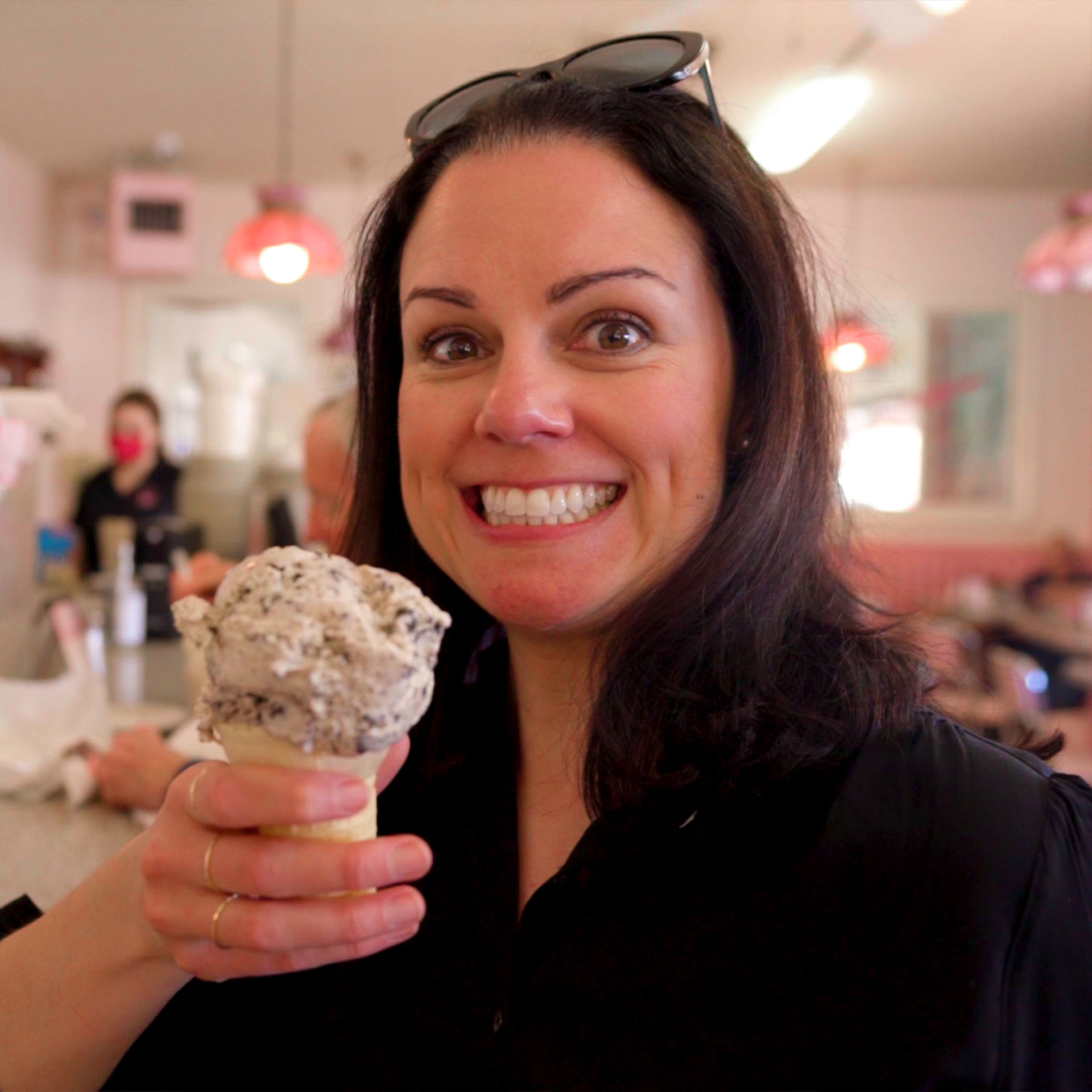 A woman wearing a black sweater and sunglasses holds an ice cream cone in a vintage diner.