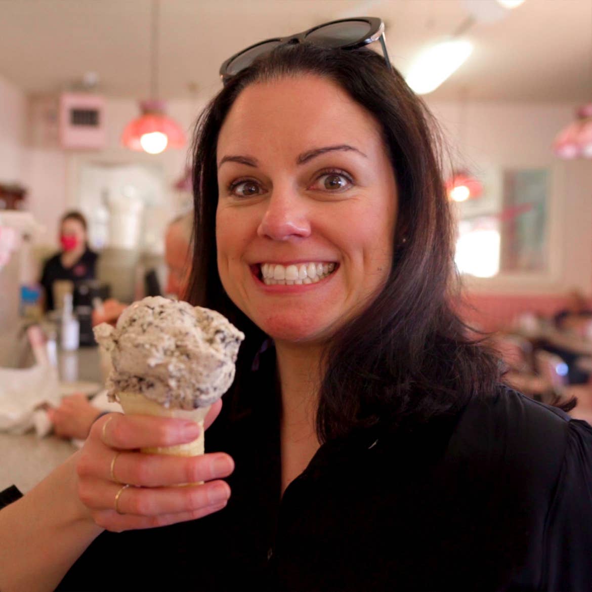 A woman wearing a black sweater and sunglasses holds an ice cream cone in a vintage diner.