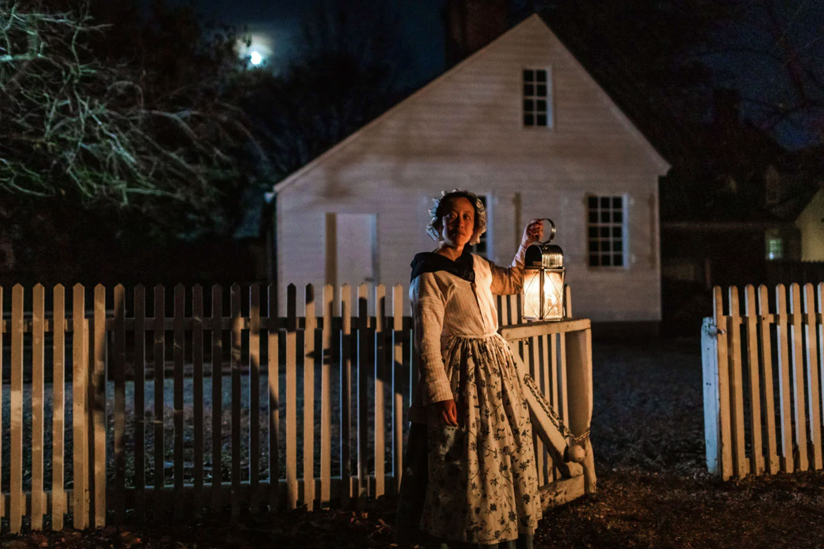 A woman in period costume stands outdoors at night holding a lantern.