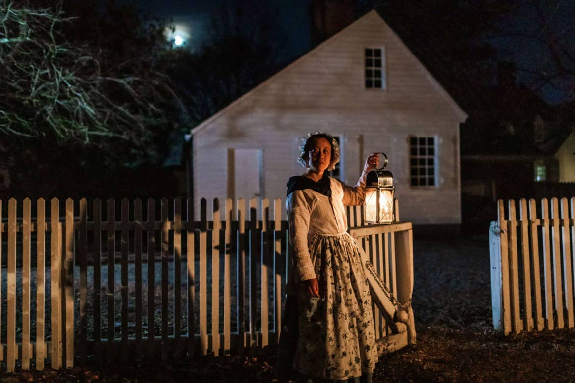A woman in period costume stands outdoors at night holding a lantern.