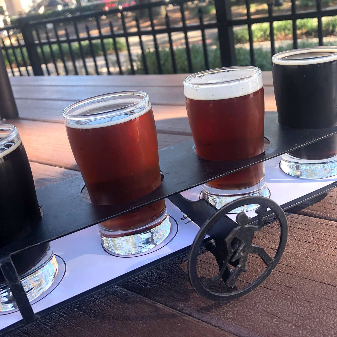 A beer flight with an iron 'Crooked Can' logo stand placed outside on a wooden plank table.