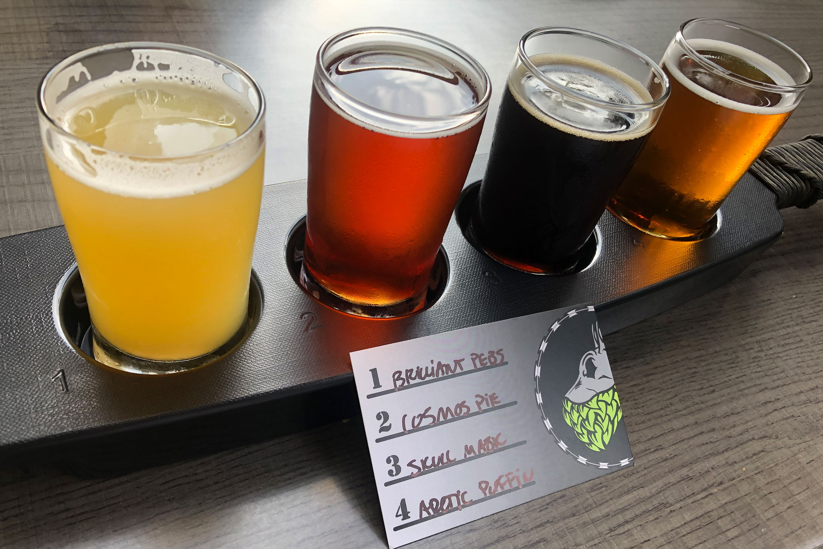 A beer flight with a label that reads, '1. Brilliant Pebs 2. Cosmos Pie 3. Skull Mask 4. Arctic Puffins' from 'Tactical Brewing Co.'