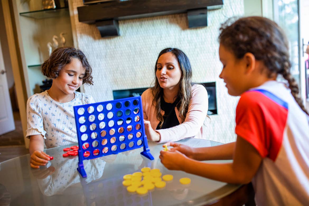 A woman watches two girls play Connect Four in a living room.
