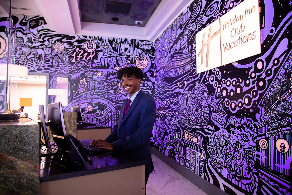 HICV Team Member, Ruben, stands in front of a mural with animated projection mapping in various colors of purple, orange, yellow and magenta featured in the lobby of our resort in New Orleans, Louisiana.