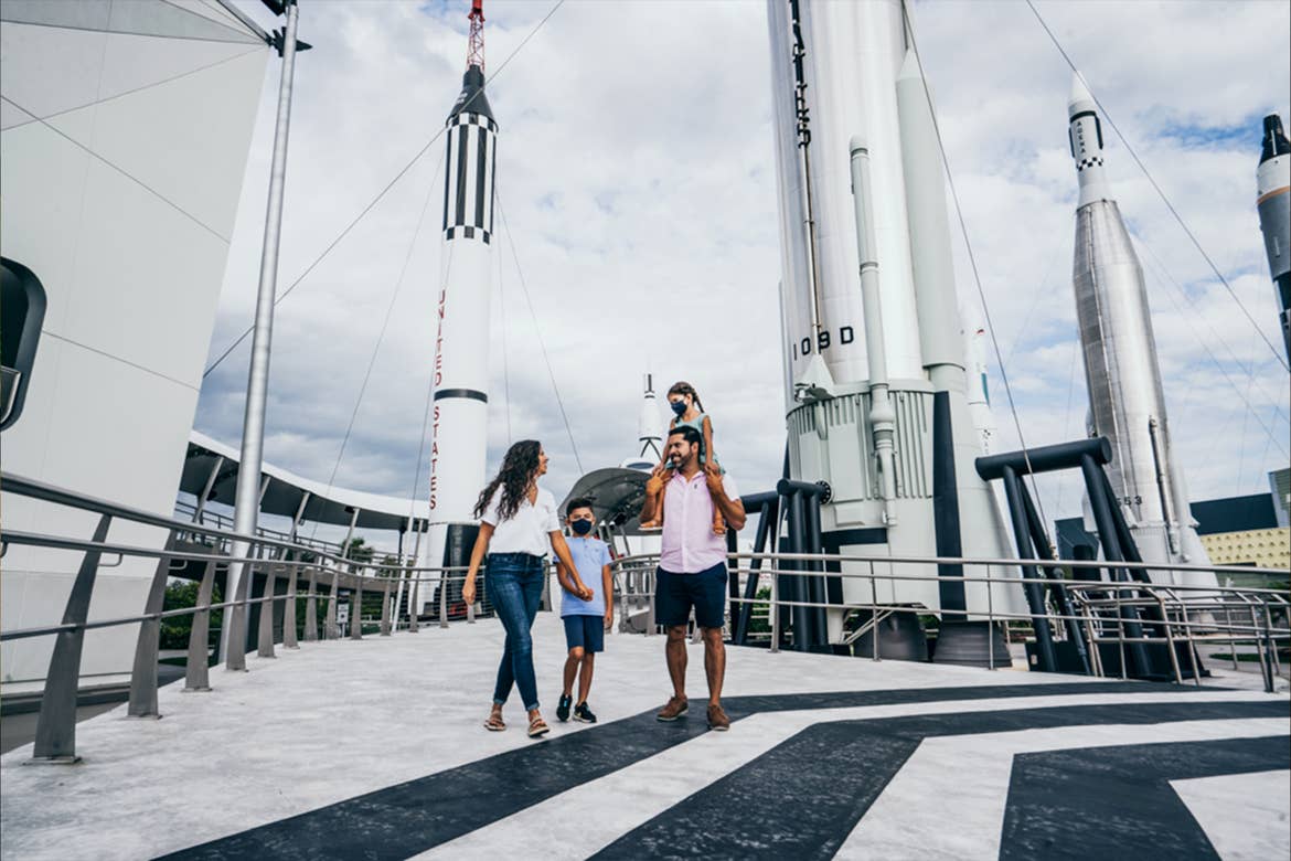 A woman in a white shirt and jeans holds hands with a young boy in a blue shirt and shorts next to a man in a pink button-up shirt and dark shorts carrying a young girl in a blue dress near a Rocket Garden.