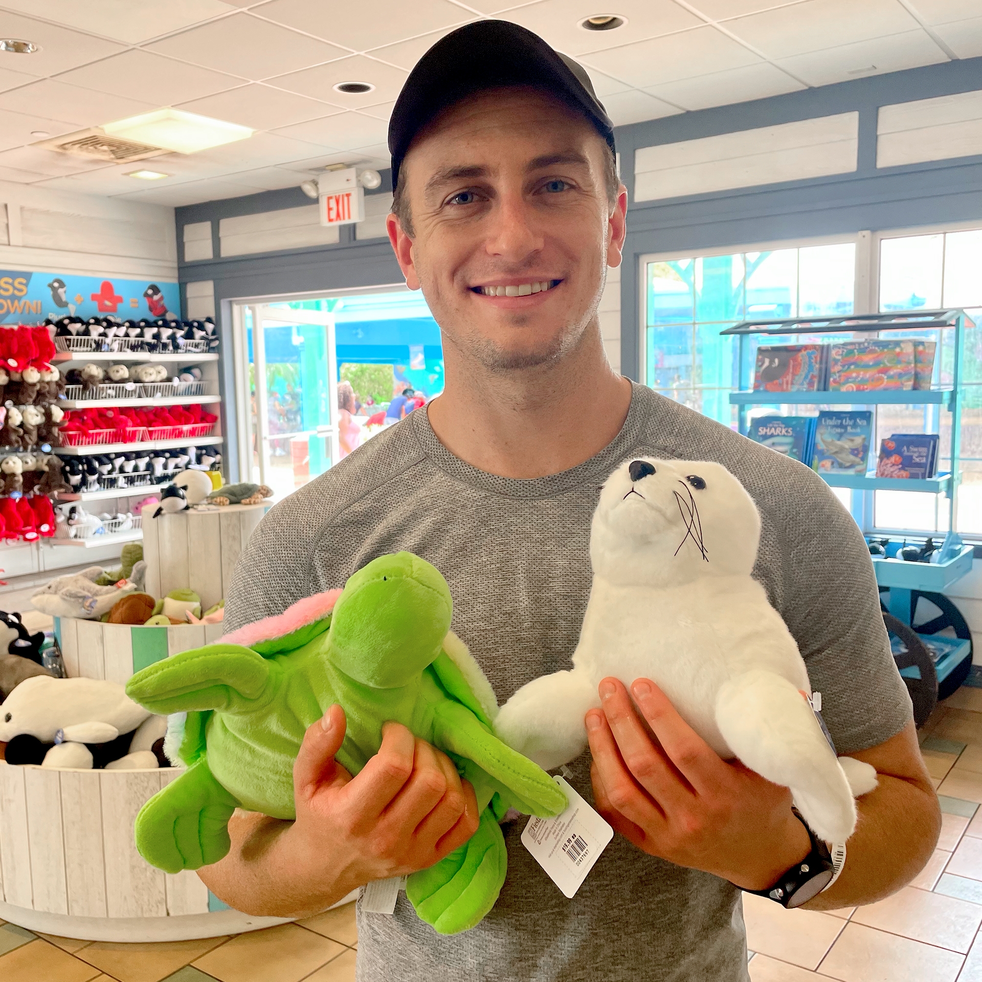 A Caucasian male wearing a grey t-shirt and black cap holds a stuffed turtle and arctic seal in a shop.