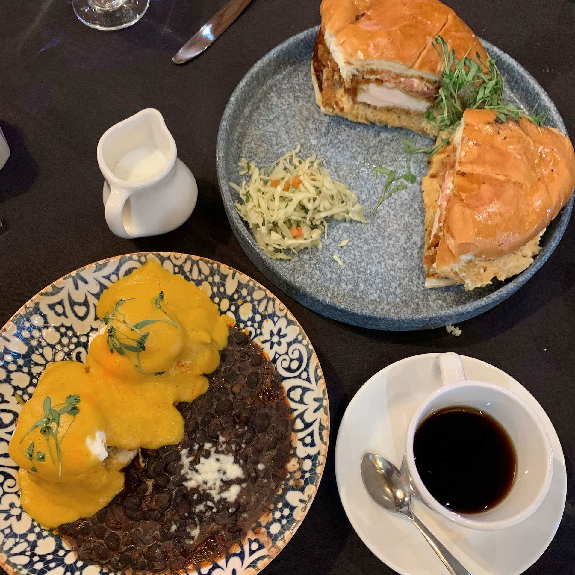 Two dining plates containing various dishes and one white saucer with a mug of coffee are placed on a dark wood dining table.