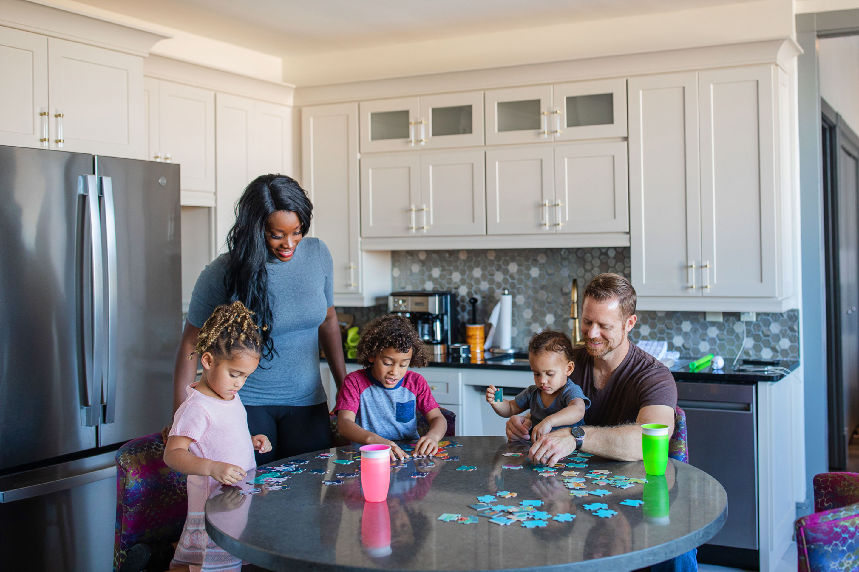From left to right: A young girl, woman, young boy, and a dad holding a toddler sit at a kitchen table working on puzzles.